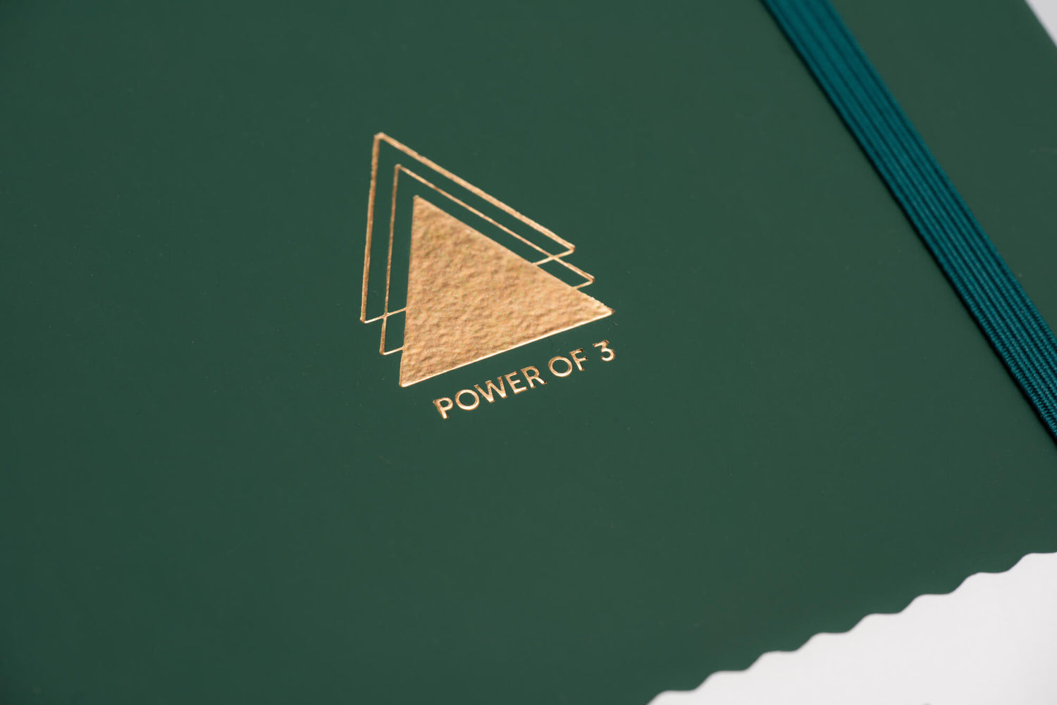 Close up image of triangle emblem from the Power of 3 goal planner, in gold foil on green journal