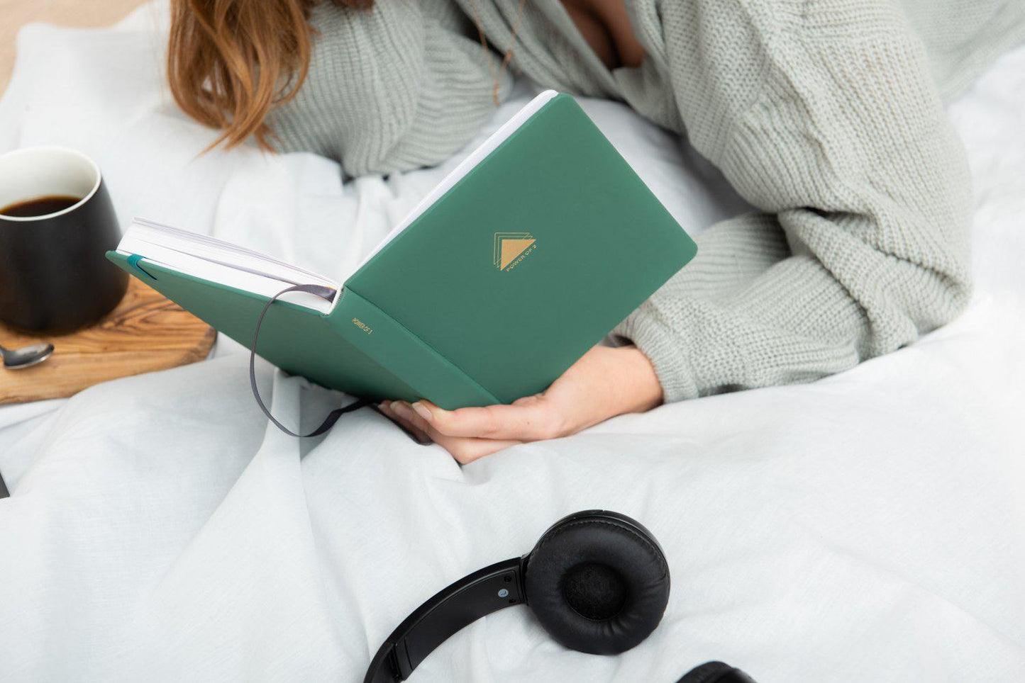 Woman lying on bed holding green power of 3 goal planner with coffee and headphones